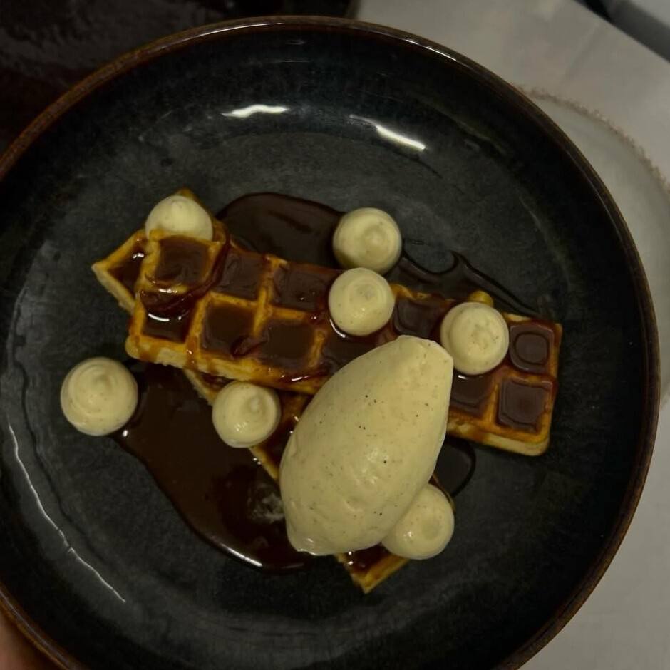 Gaufre nappée de sauce Suzette, points de crème mousseline et quenelle de glace vanille, dans une assiette creuse sombre.