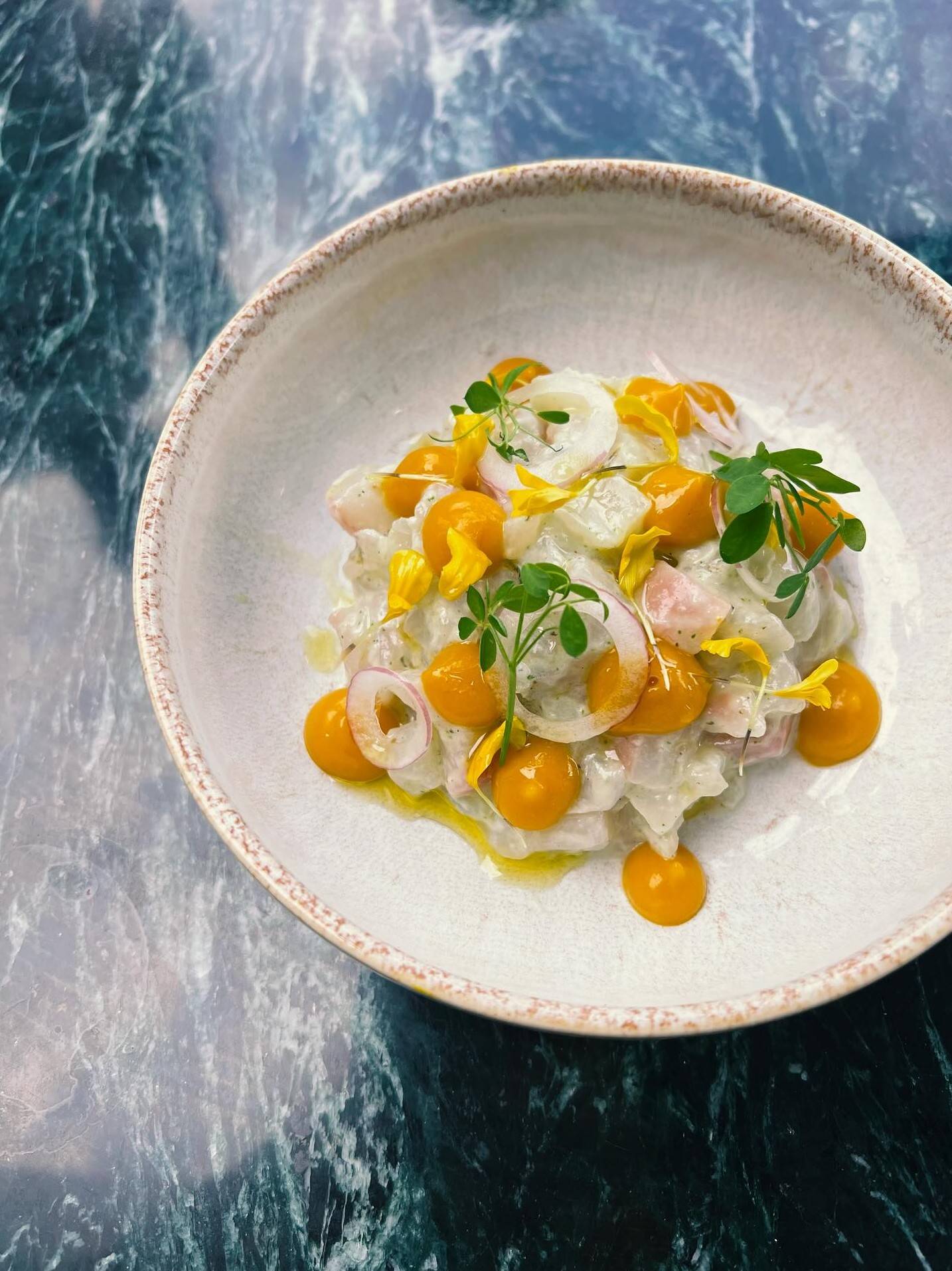 Crudo de poisson vu du dessus, dans une assiette creuse blanche au bord moucheté : points de gel orange, rondelles d’oignon rouge, pétales jaunes et jeunes pousses, sur marbre vert.