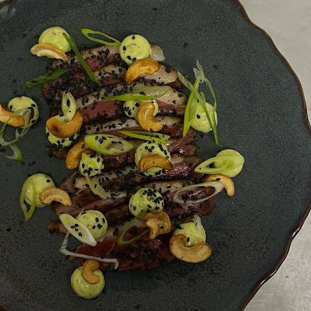 Tataki de magret vu du dessus : tranches de magret, noix de cajou et points de mayonnaise à l’estragon, sur une assiette sombre.